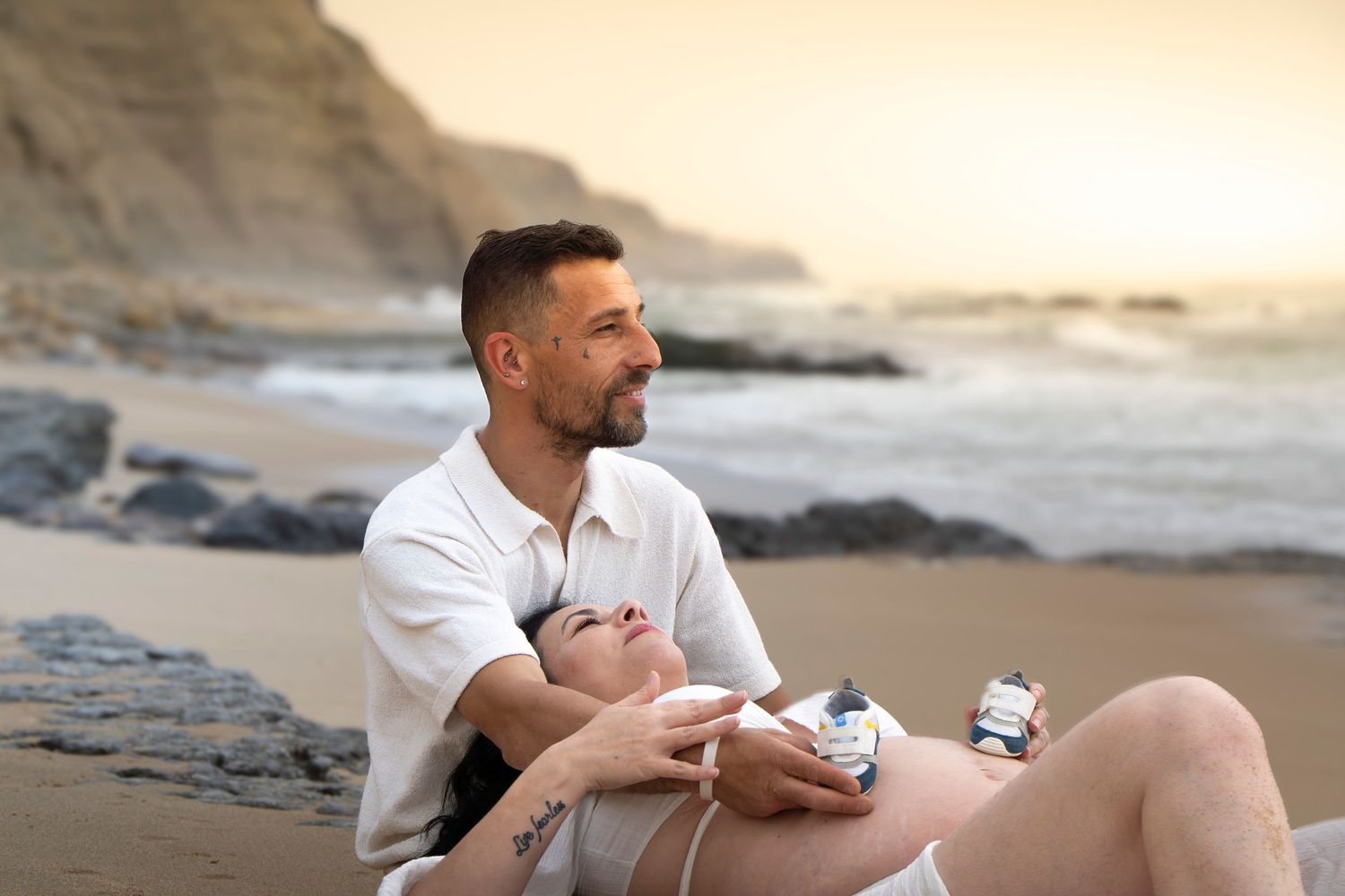 Couple relaxing on a beach at sunset, pregnant woman resting on mans lap near ocean cliffs.