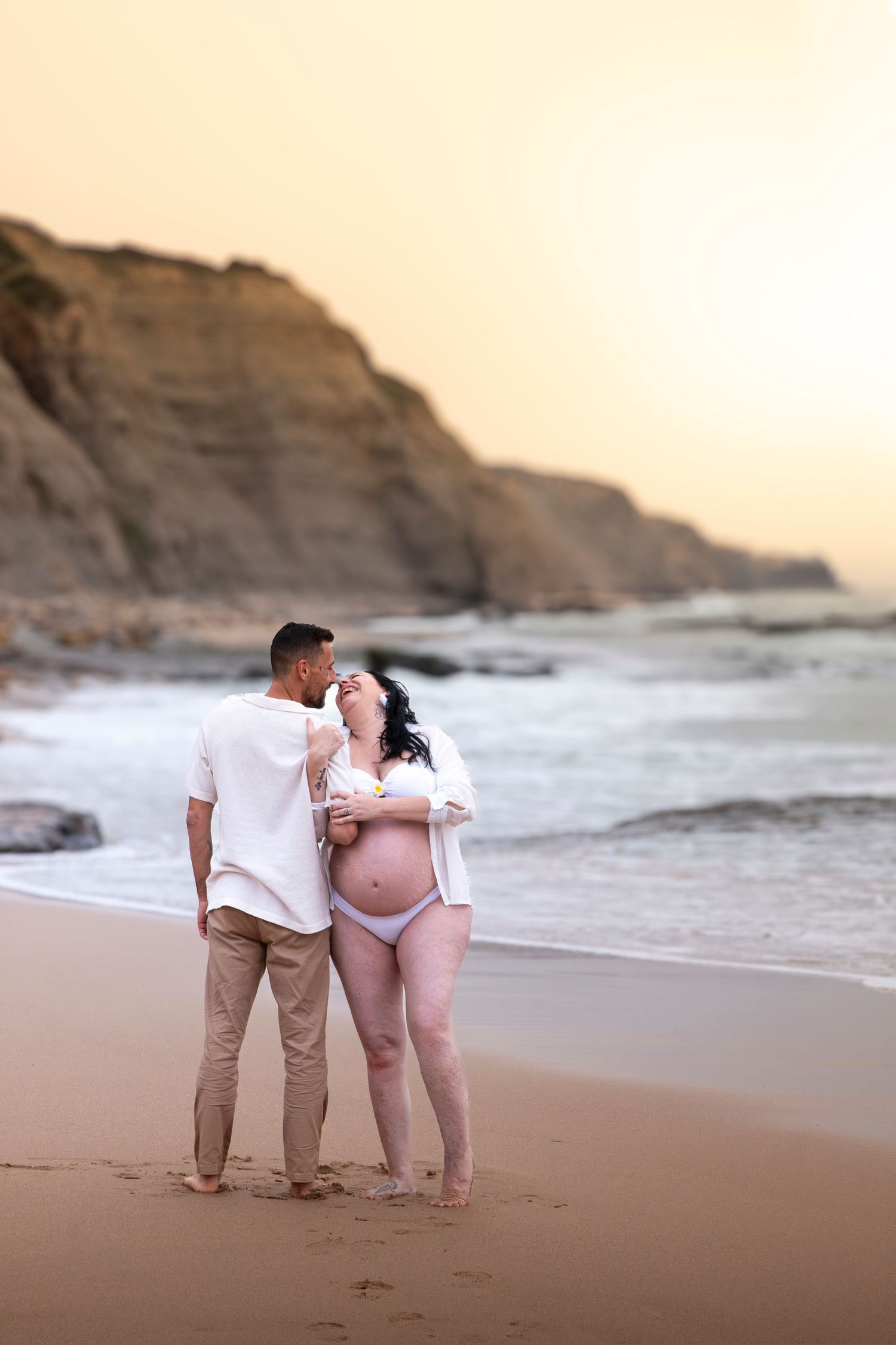 Pregnant couple embracing on a sandy beach at sunset with rocky cliffs and ocean waves in the background.