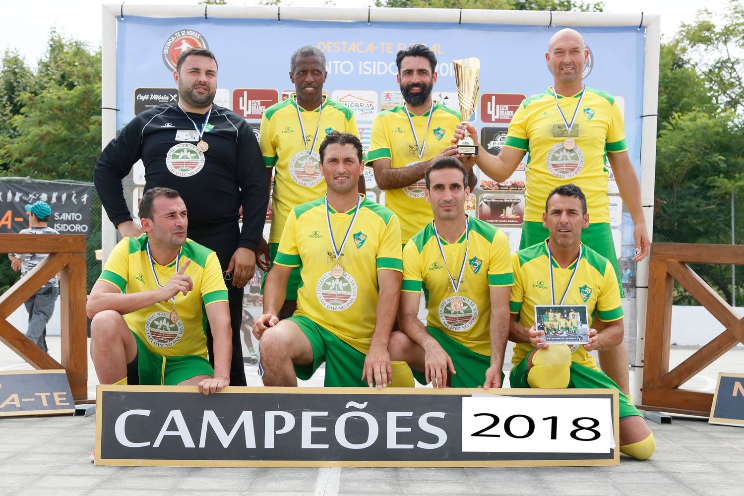 Soccer team poses with championship trophy and yellow uniforms displaying 'Campeoes 2018' banner.