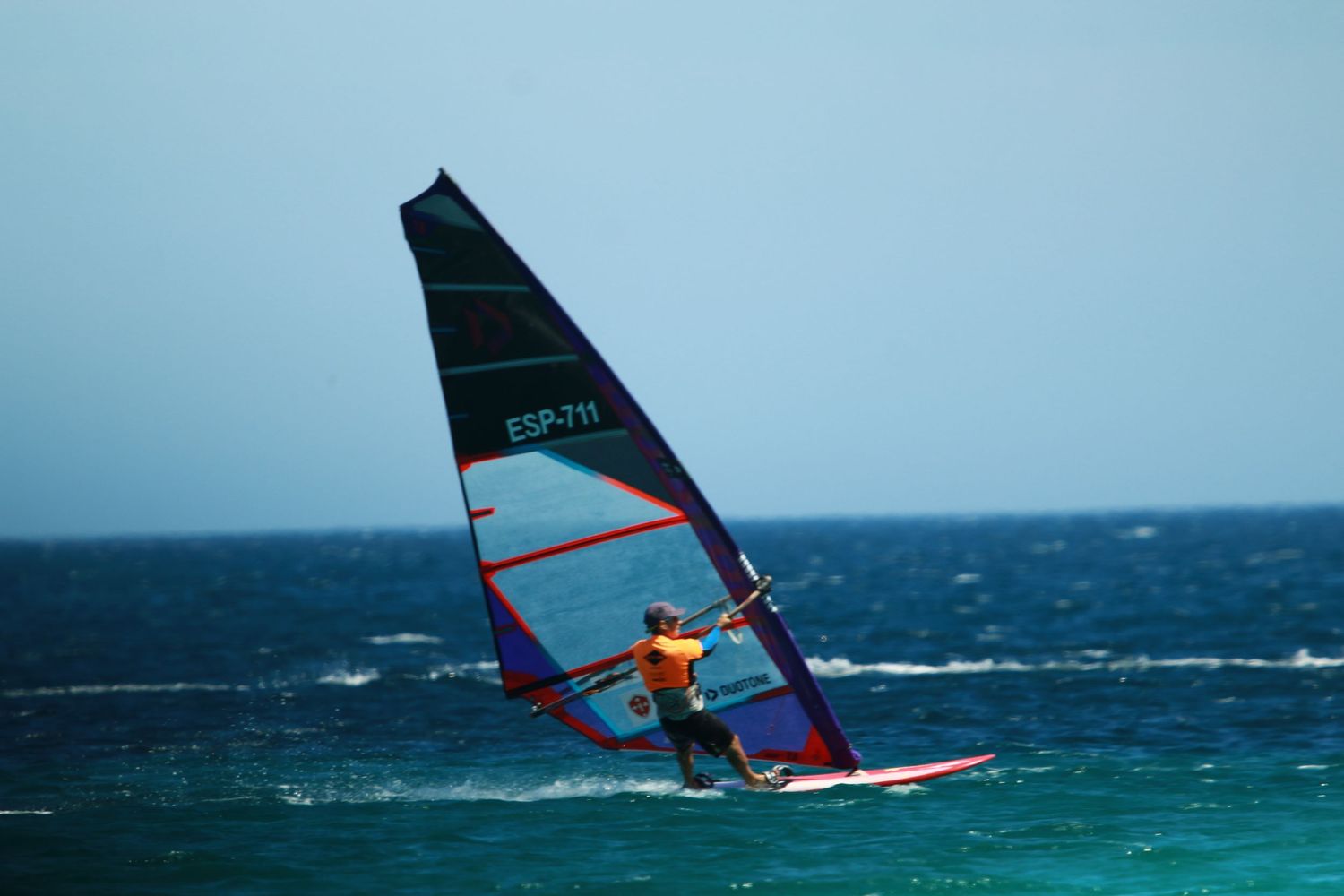Solo windsurfer navigating blue ocean waters with dark sail against clear sky.