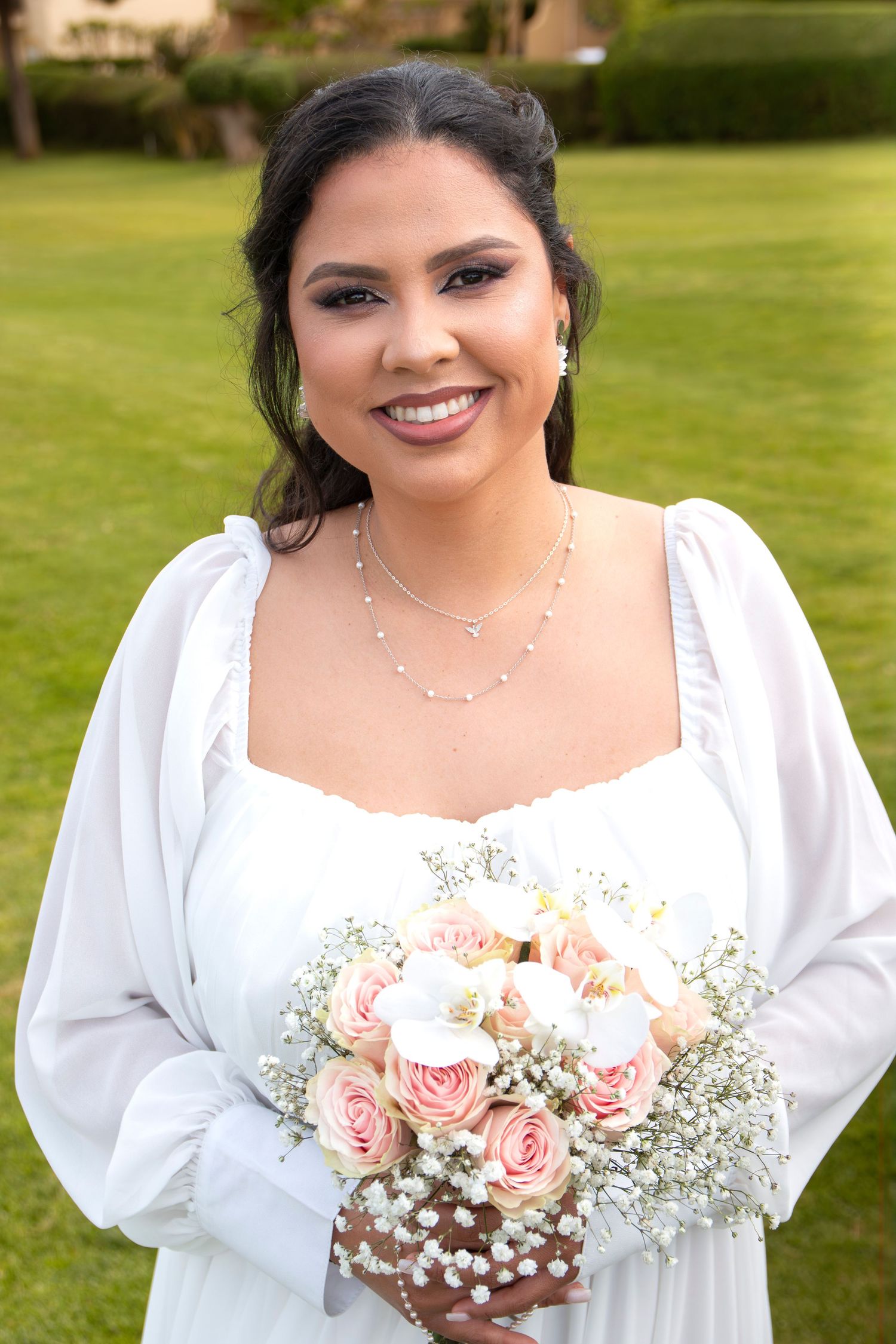 Bride in white dress holding romantic pink roses and baby's breath bouquet on a lush green lawn.