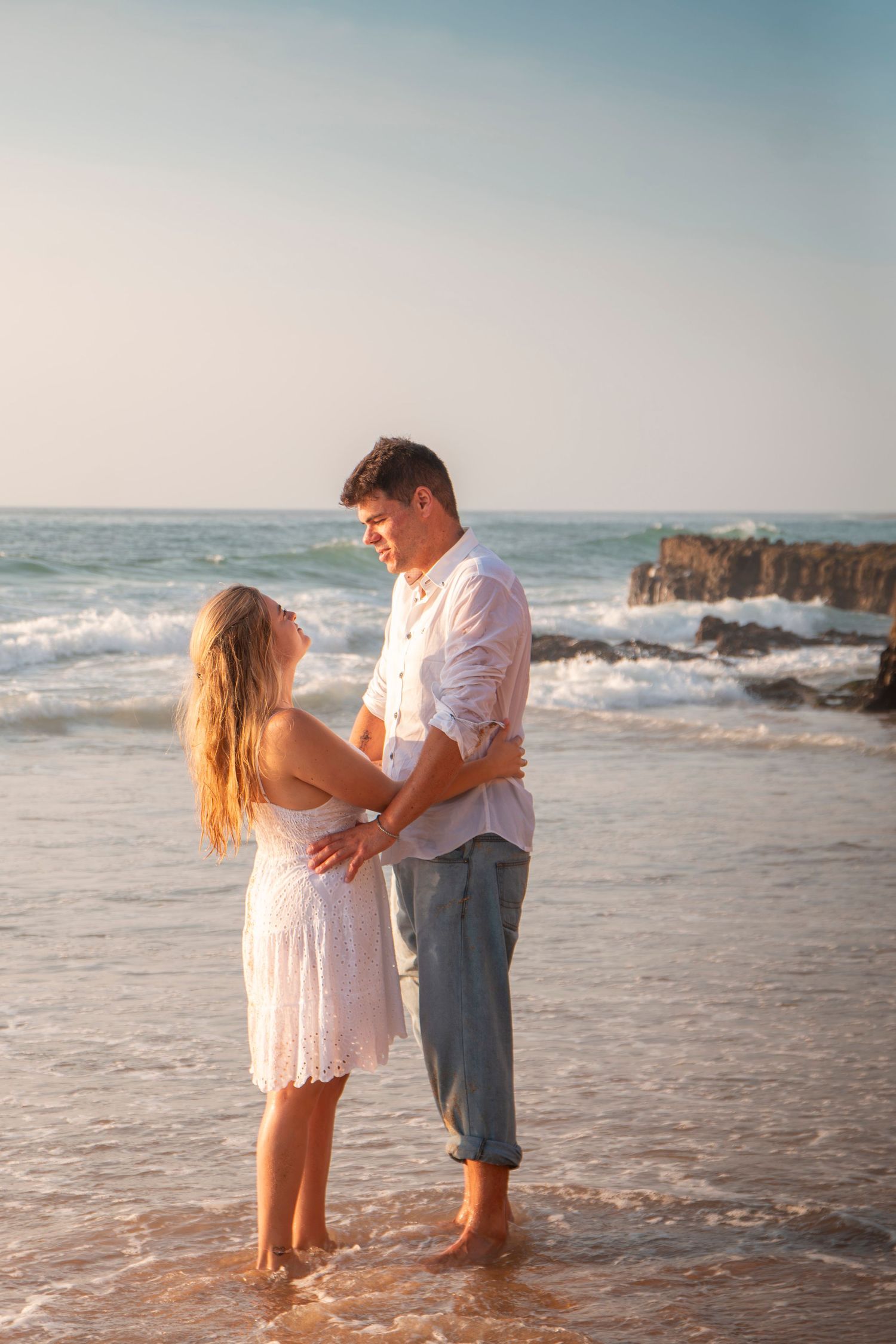 Couple shares intimate moment on sandy beach with waves crashing against rocks in background during sunset.