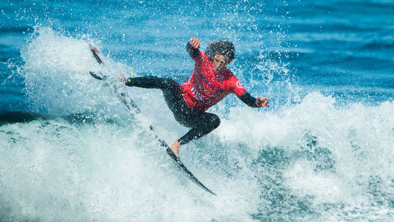 A surfer in a red wetsuit performs an aerial maneuver on a breaking ocean wave with splashing water around them.