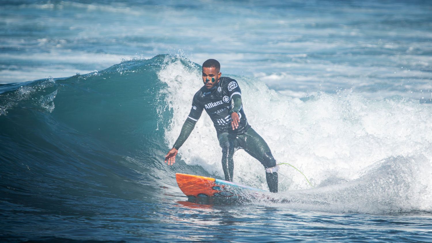 A surfer in a wetsuit rides an ocean wave on an orange surfboard against blue water and white spray.