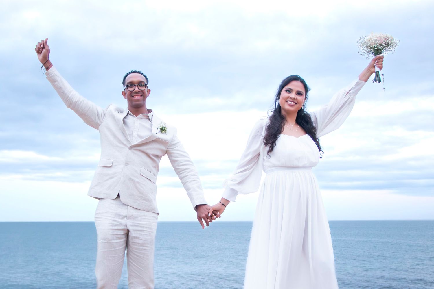 Couple in white outfits walking and posing playfully on a beach with ocean and cloudy sky in the background.