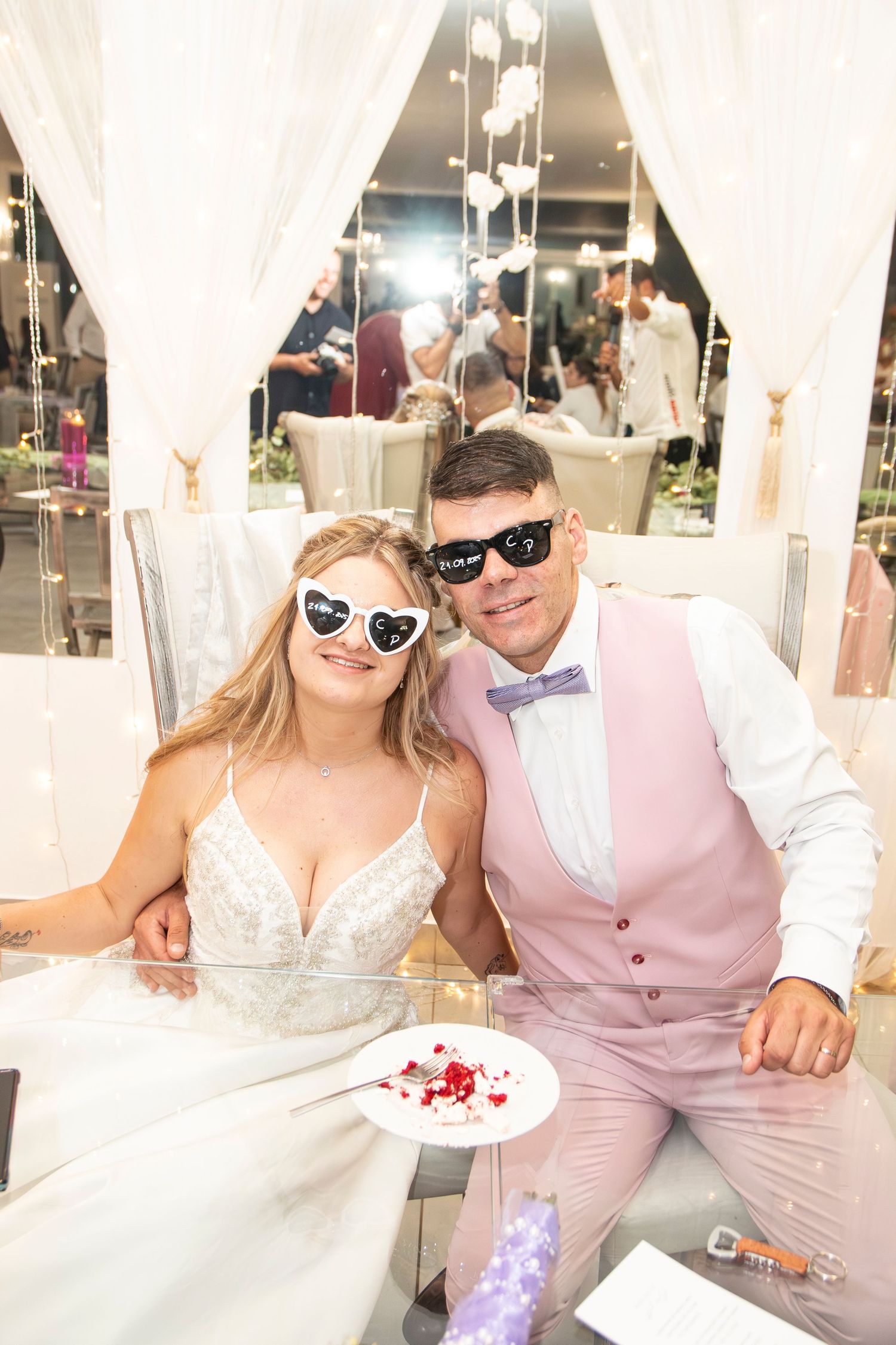 Bride and groom wearing novelty heart sunglasses smile together at their wedding reception celebration.