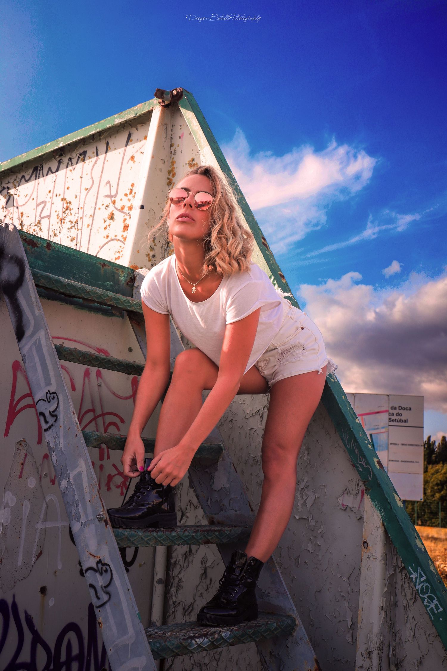Person in white t-shirt and shorts tying shoelaces on concrete steps with graffiti under bright blue sky.