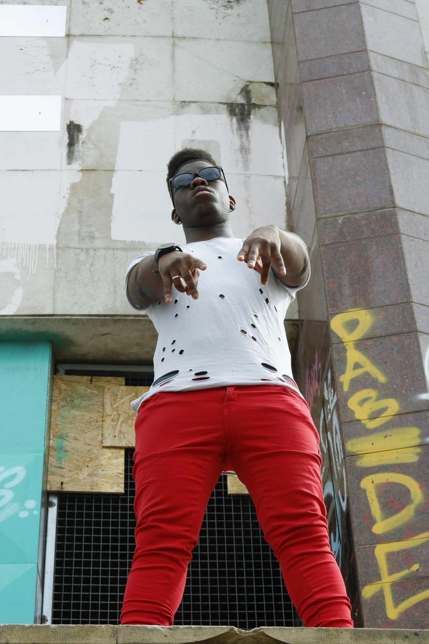 Person in a white distressed t-shirt and red pants stands on concrete steps with graffiti in the background.