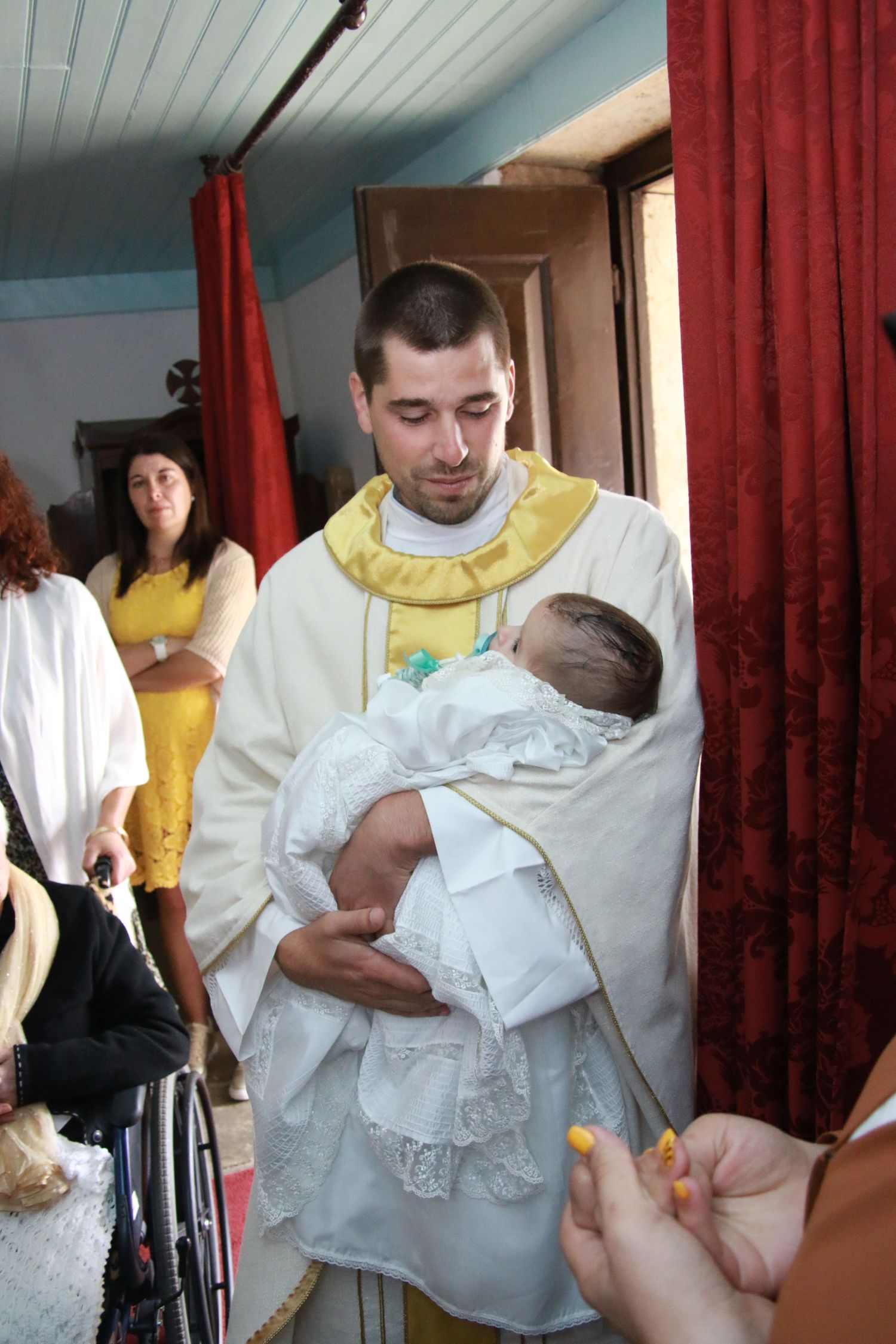 Religious ceremony with clergy member in yellow and white vestments holding a baby in white baptismal gown.