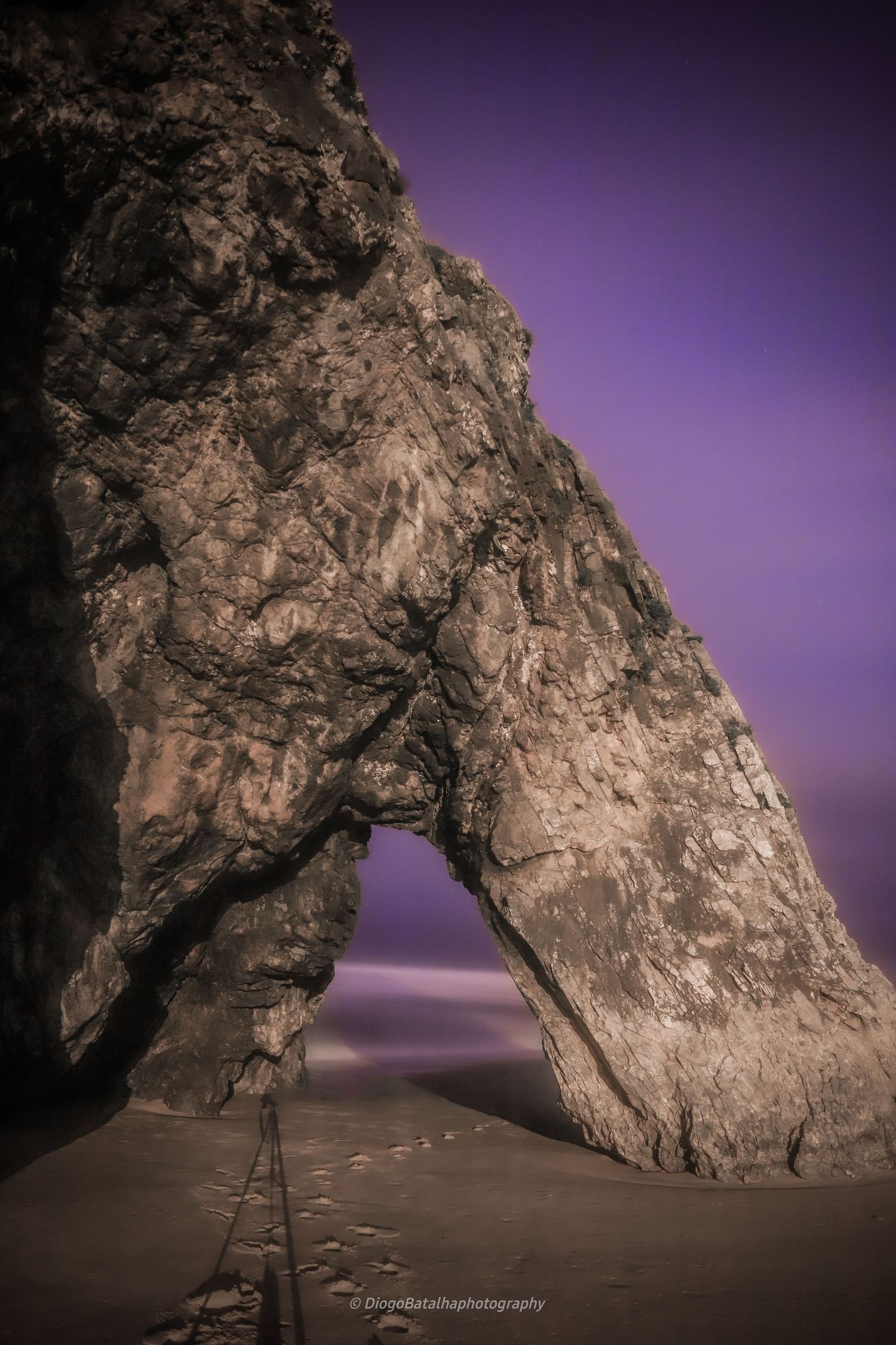 A dramatic natural rock arch formation on a beach at twilight with purple sky and sandy foreground.