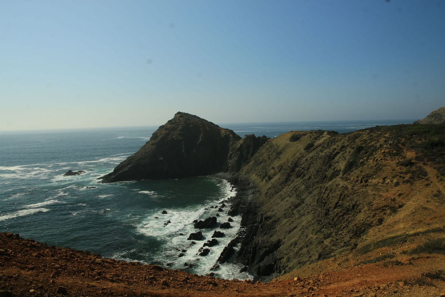 Rugged California coastline with dramatic cliffs and crashing waves against rocky shores under clear blue skies.