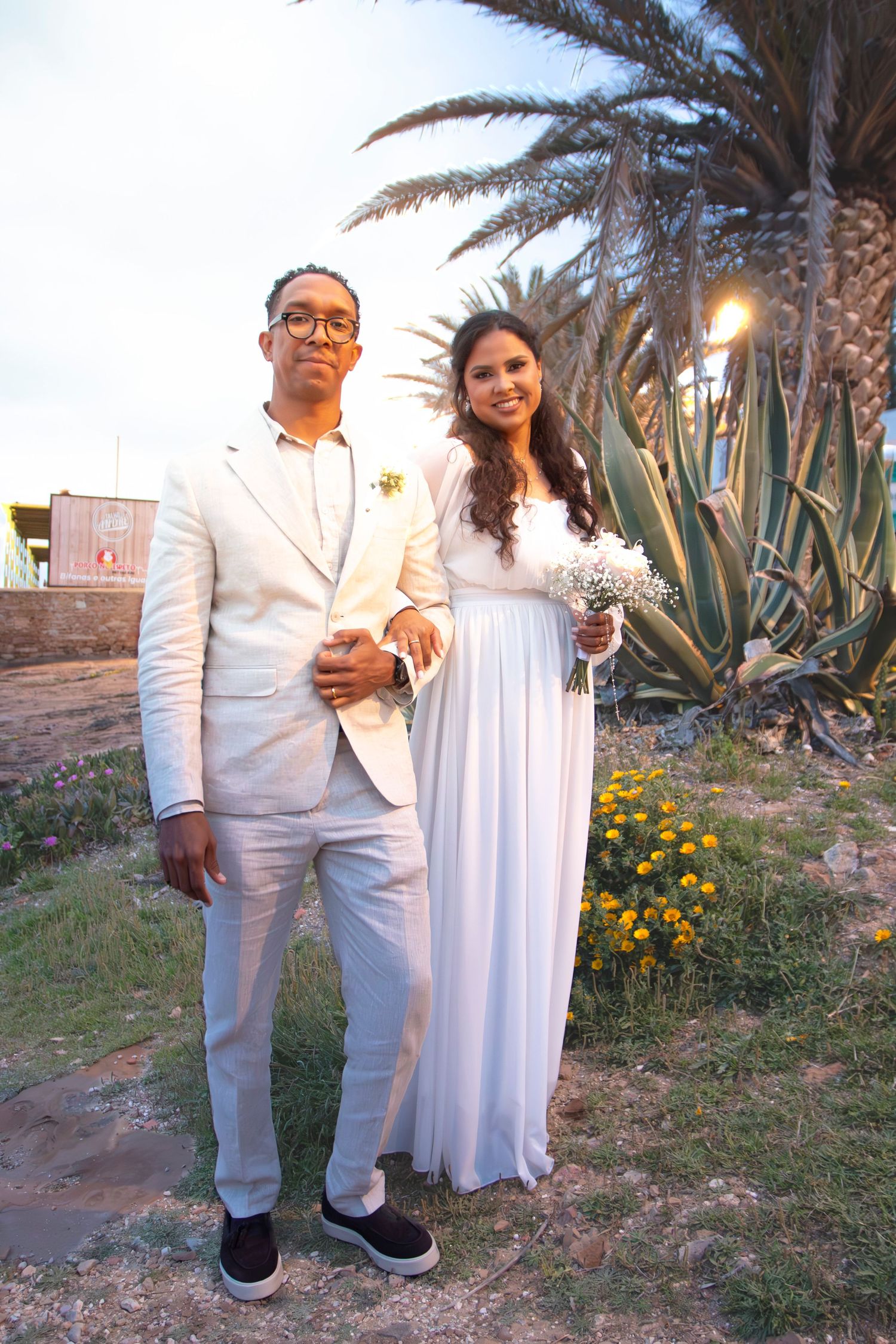 A couple in coordinated white outfits poses together in a garden setting with palm trees and yellow flowers at dusk.