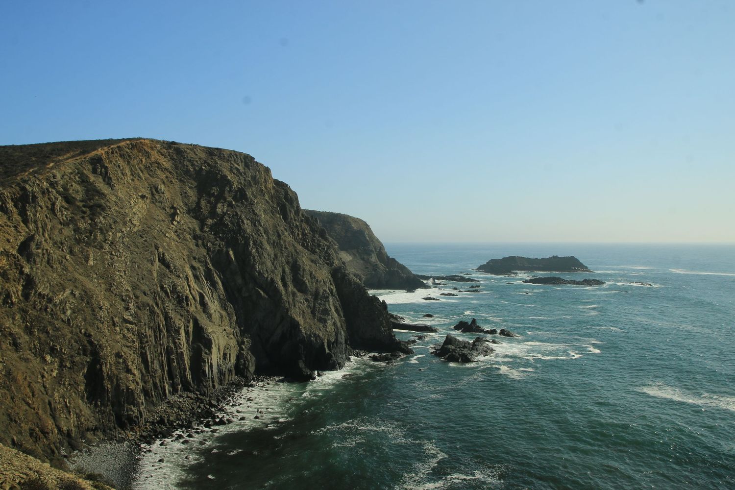 Rugged coastal cliffs rise above crashing waves along the Pacific Ocean with offshore rocks visible in the distance.