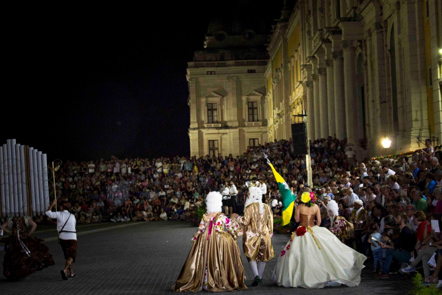 Performers in period costumes entertain a large nighttime crowd gathered outside an illuminated historic building.