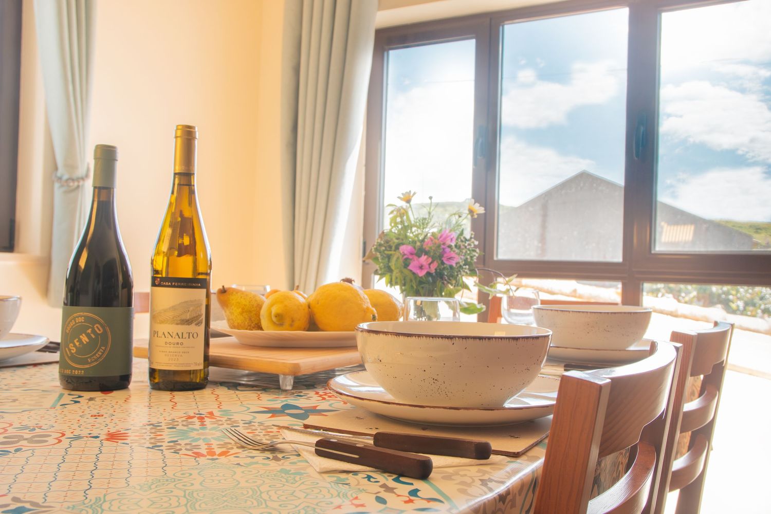 Dining table set with wine bottles and soup bowl by window overlooking mountain view with sunlight streaming in.