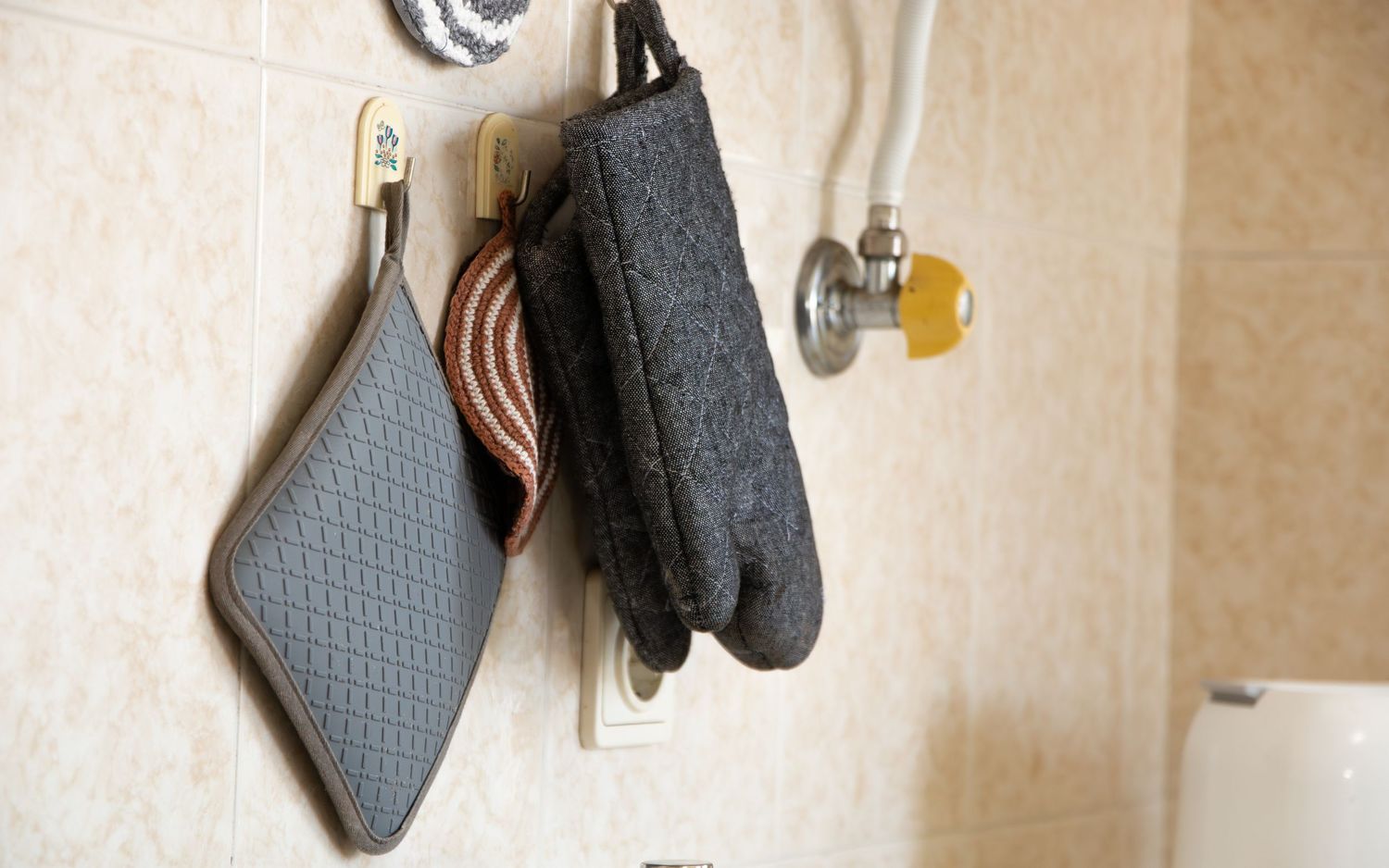 Gray and black textured oven mitts hanging from metal hooks on a beige wall.