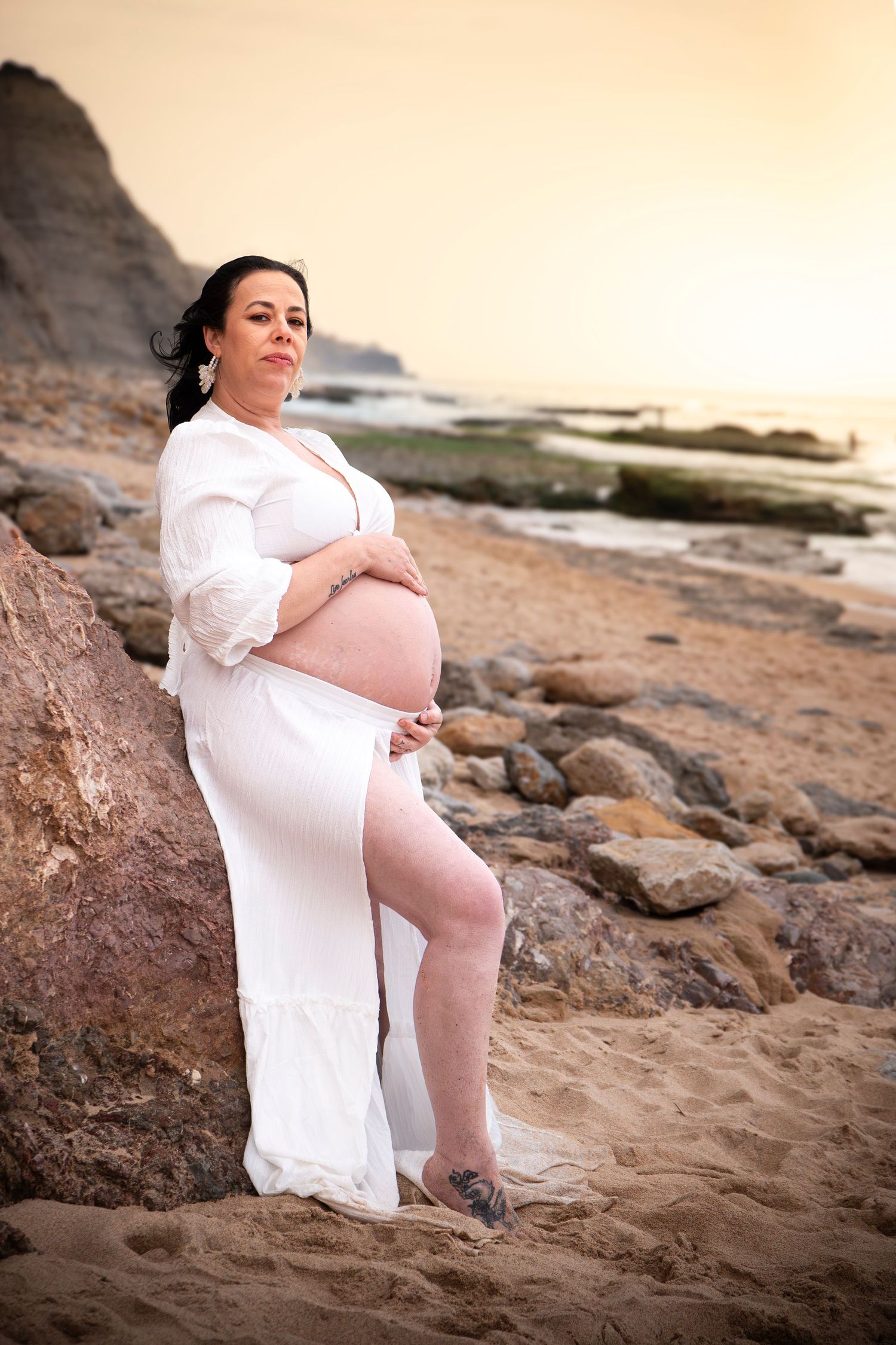 Pregnant woman in white dress cradling baby bump on rocky beach at sunset.