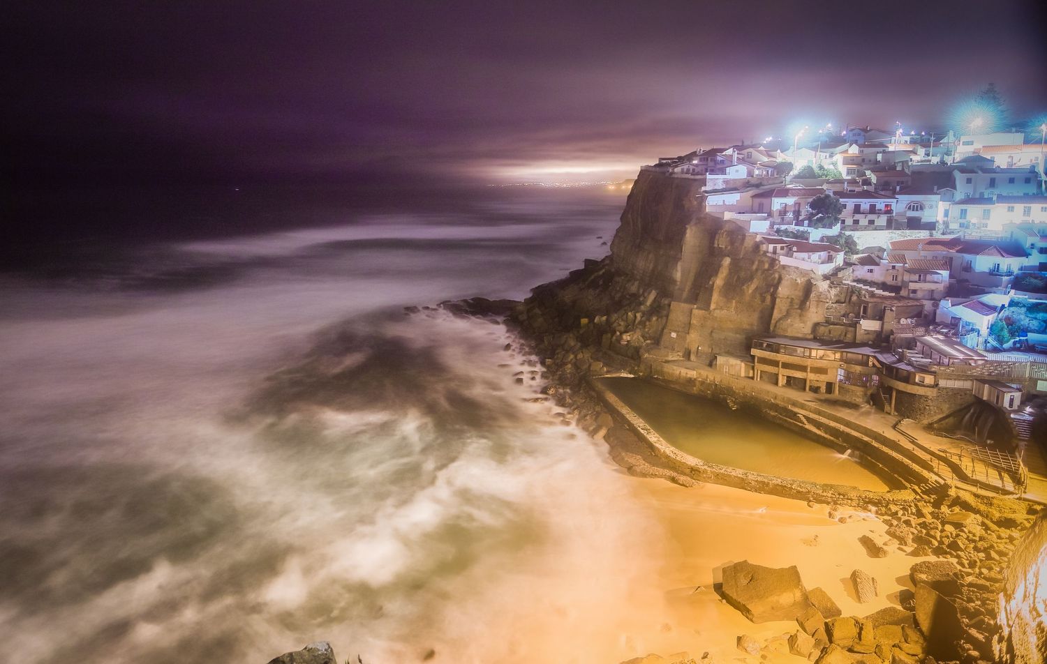 Beautiful aerial view of illuminated cliff-side town Azenhas do Mar in Portugal at night with misty ocean waves.