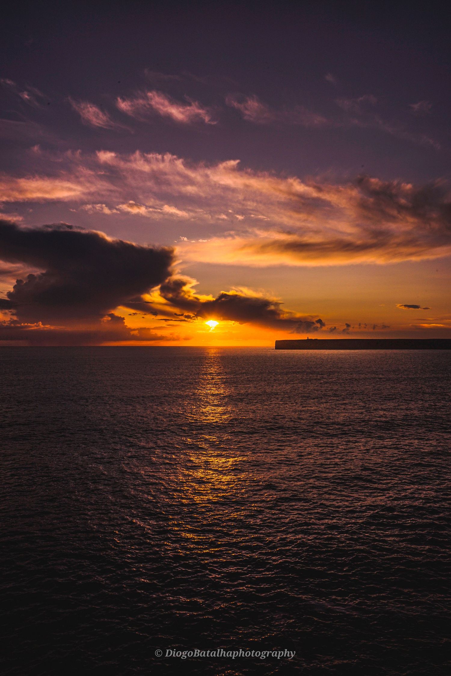 Dramatic orange sunset over dark ocean waters with silhouetted clouds and coastline reflecting golden light on waves.