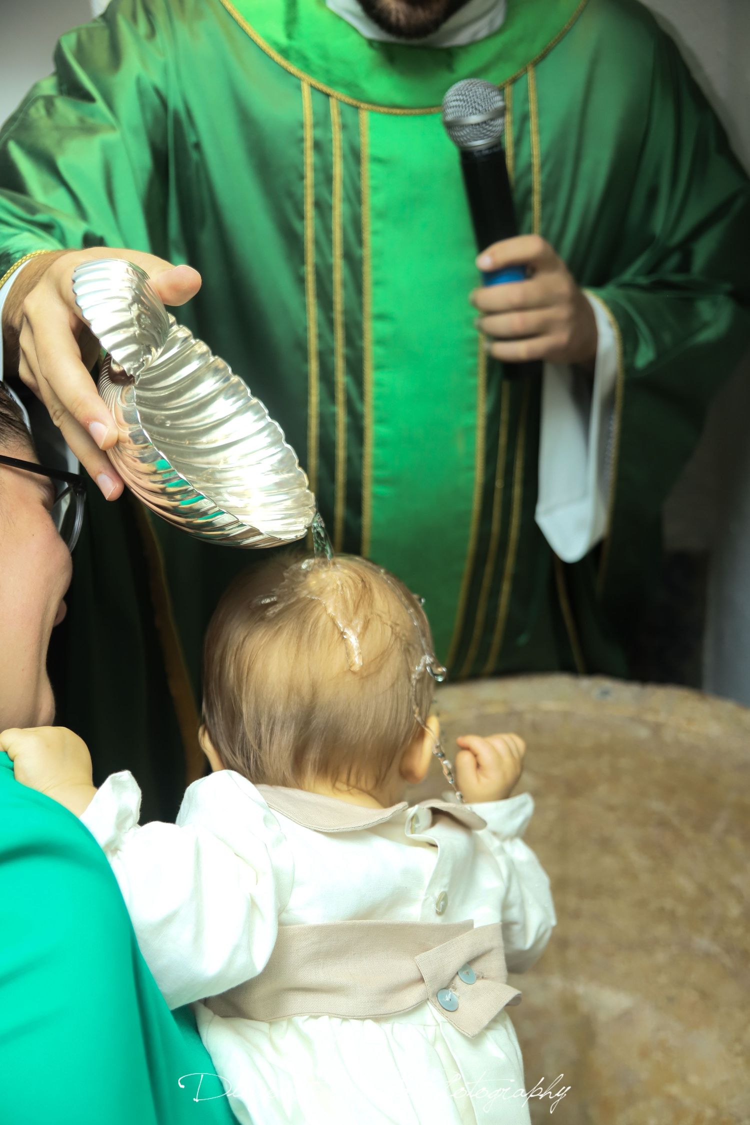 Baby in white christening gown being held by person wearing green ceremonial robes.
