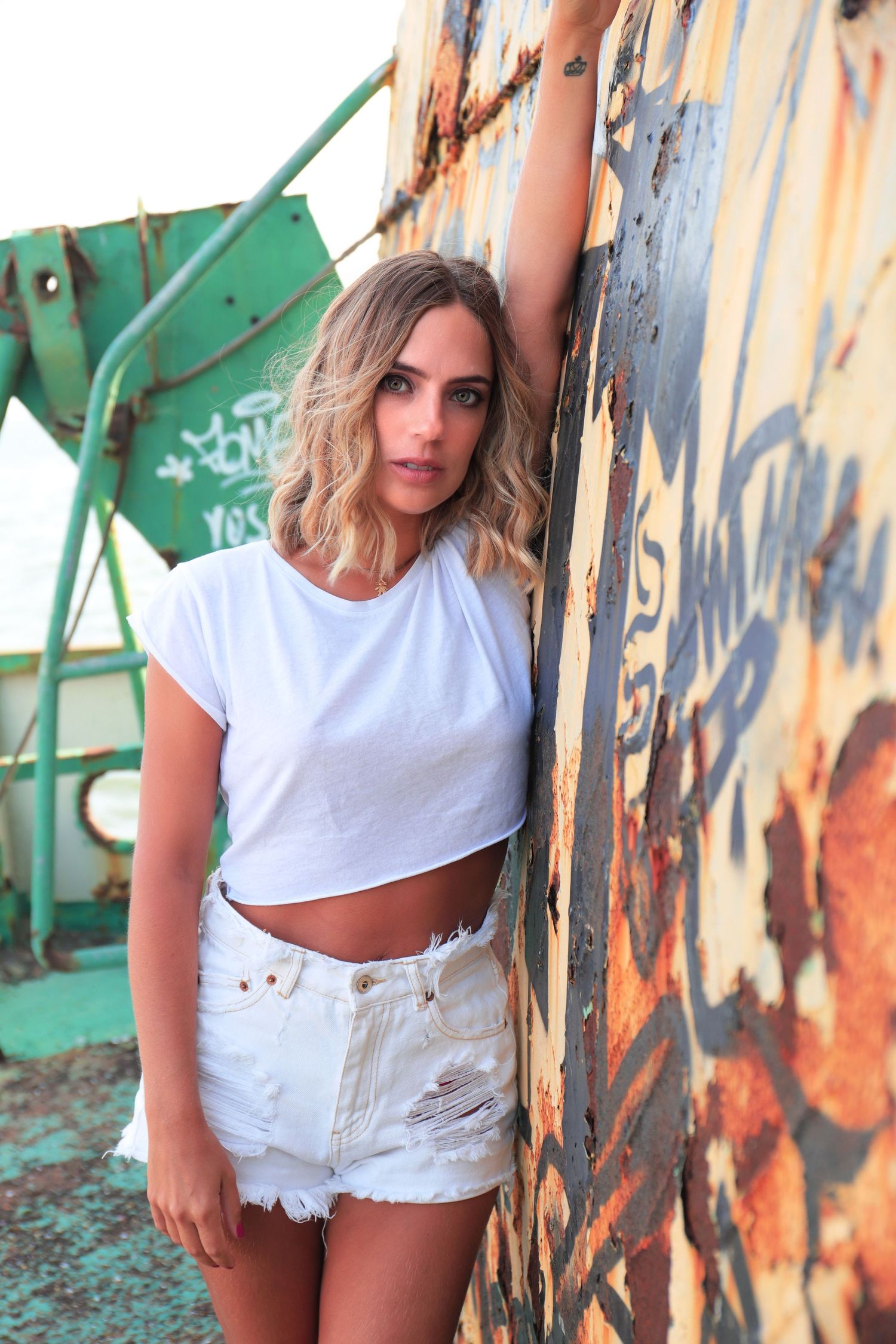A casual summer outfit with white crop top and distressed denim shorts against a graffiti wall and turquoise background.