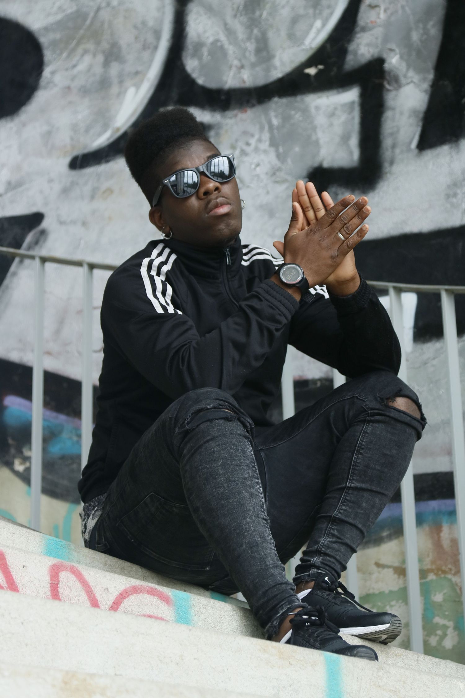 Cool streetwear style with black Adidas jacket, ripped jeans, sneakers and sunglasses against graffiti wall.