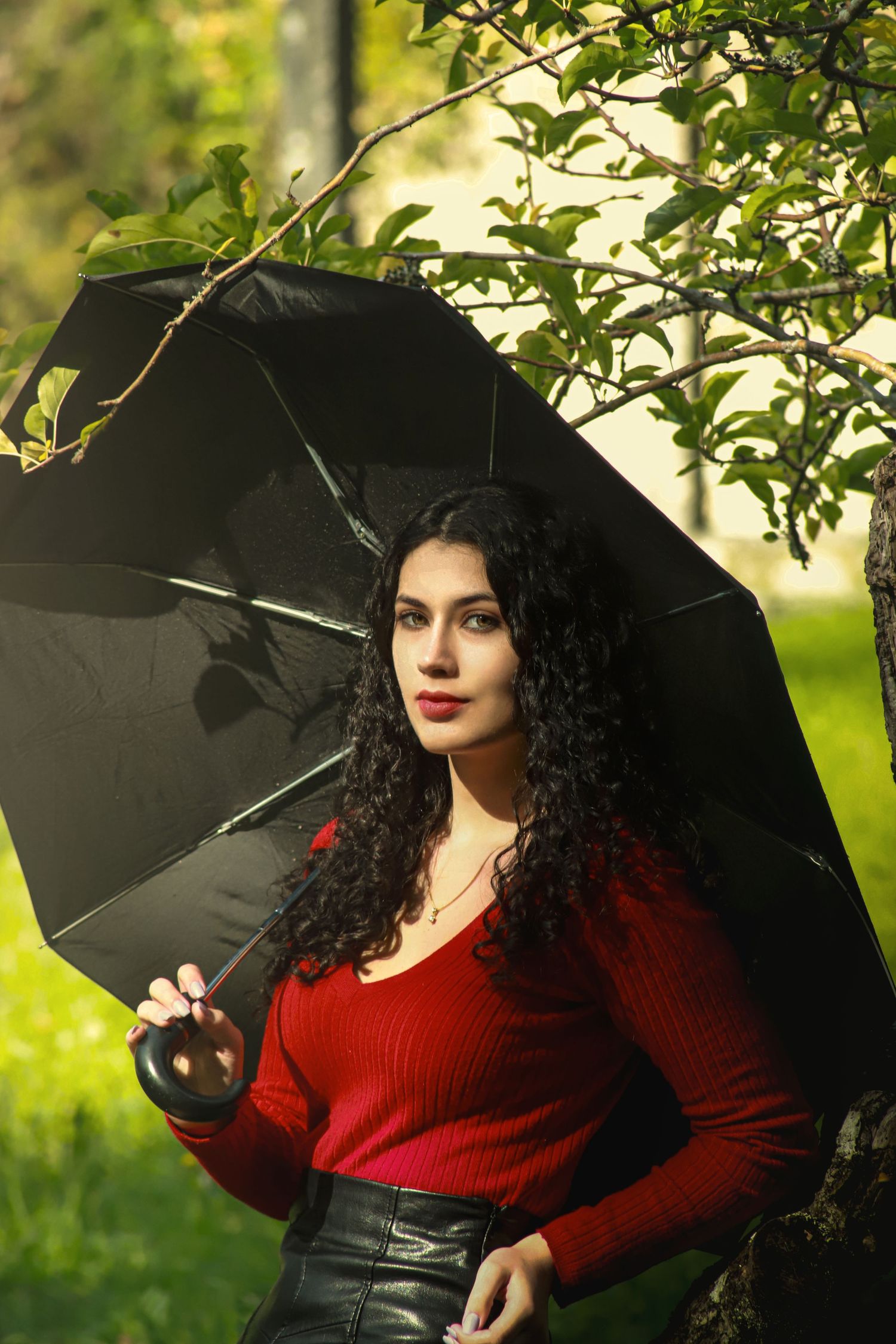 Figure holding a black umbrella while wearing a red sweater and leather skirt stands among green foliage in natural lighting.