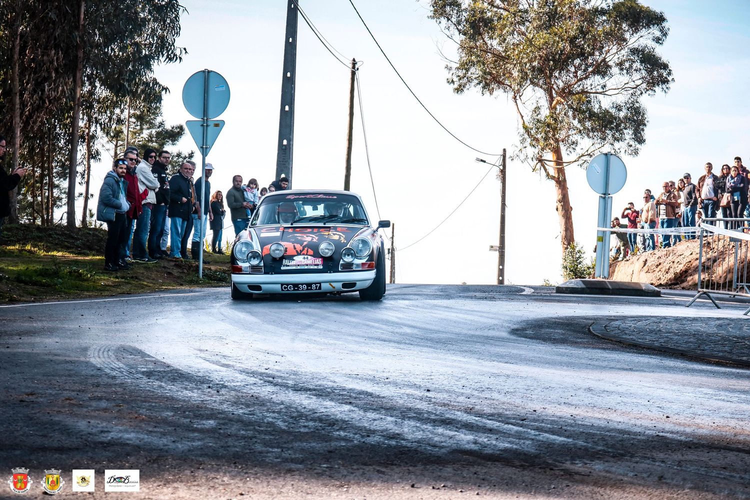 Rally car driving on winding mountain road with spectators watching from the sidelines.