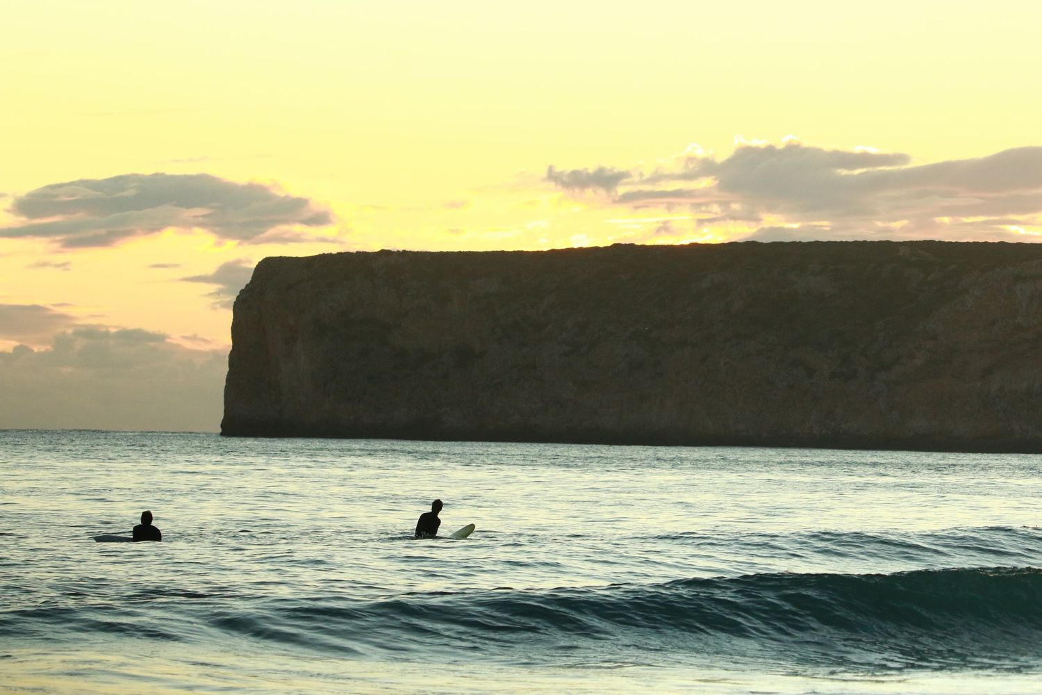 Silhouettes of surfers waiting in water at sunset with dramatic cliff backdrop.