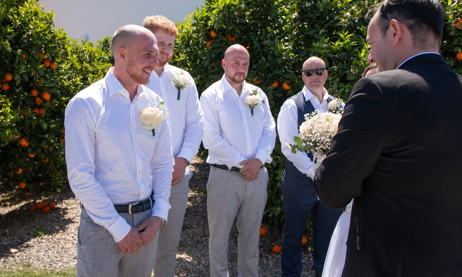 Groomsmen in white shirts and formal wear gather during outdoor ceremony.