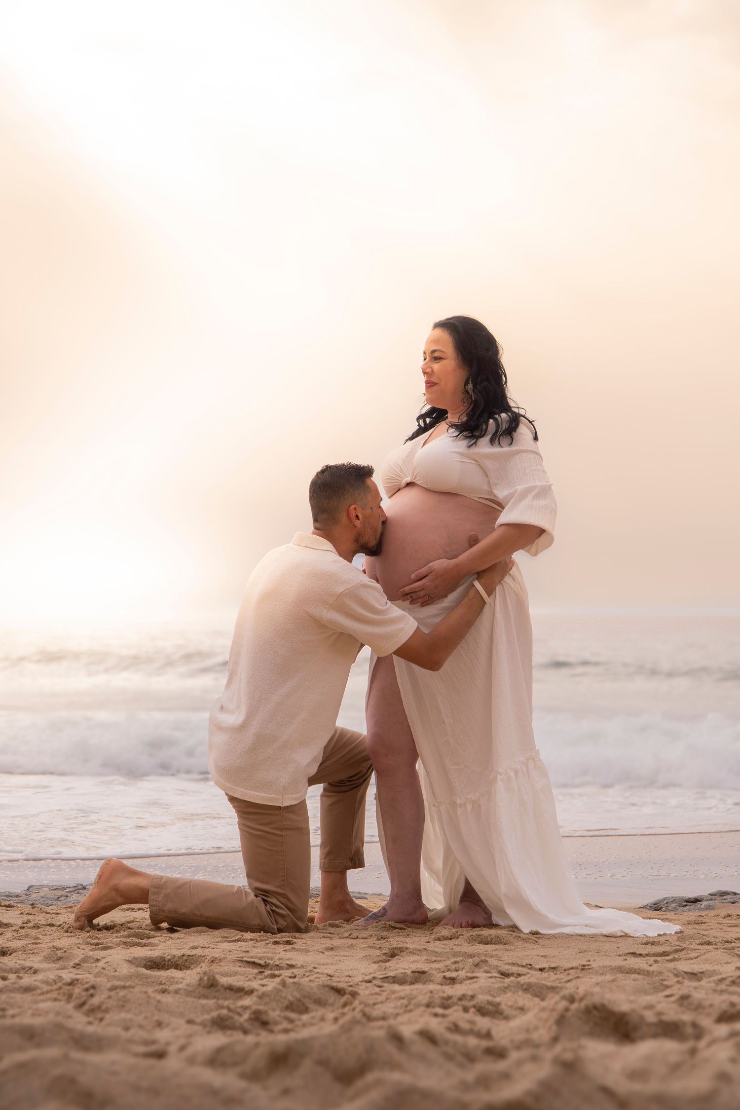 A man kneels kissing pregnant woman's belly on a sandy beach at sunset, both dressed in white.