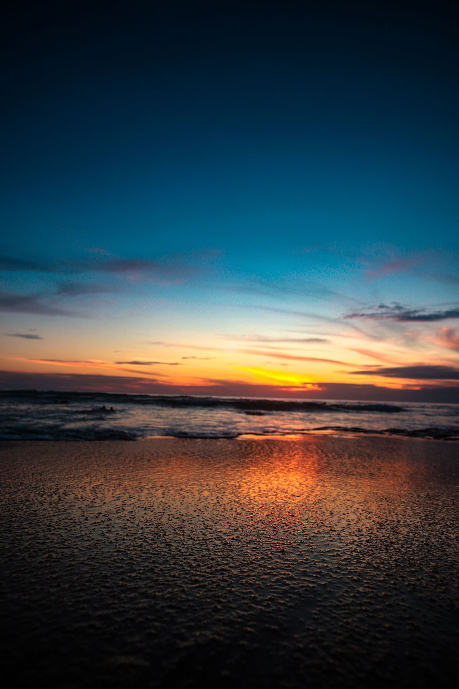 Vibrant orange sunset reflects on wet beach sand as waves roll in under a deep blue and purple twilight sky.