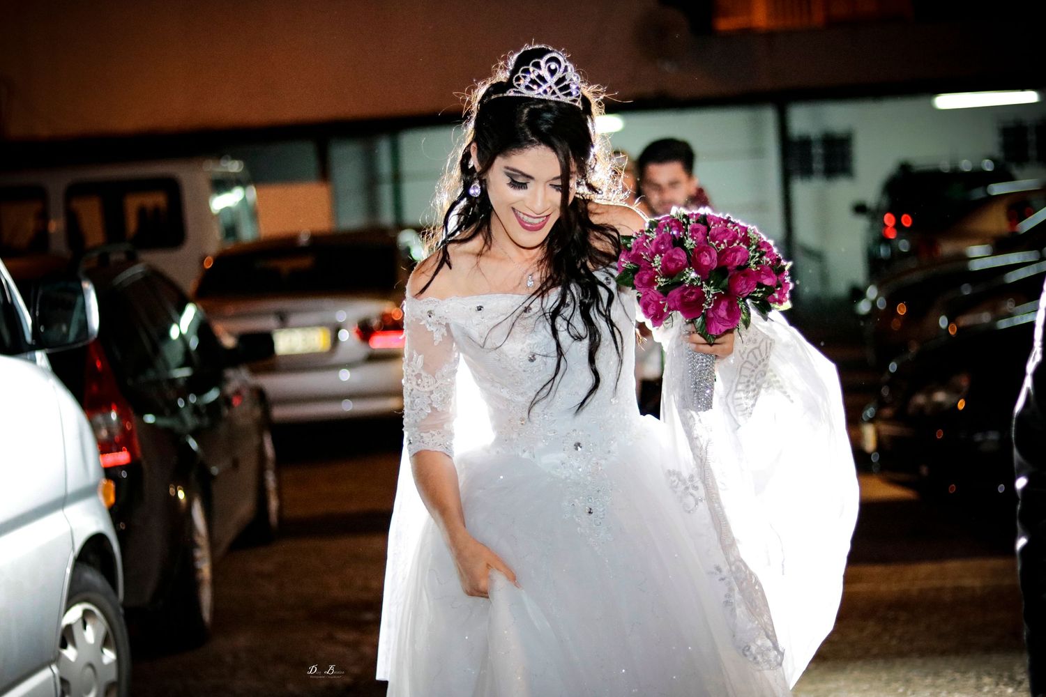 Bride in white gown with tiara holding purple bouquet in evening setting.