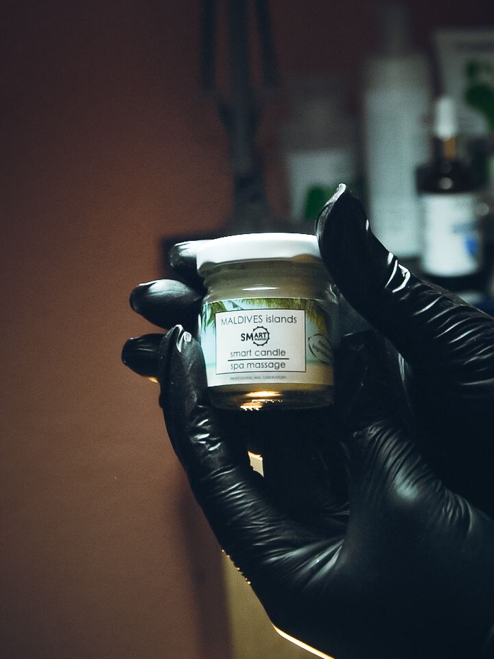Black gloved hands holding a small glass jar with cream product against dark background.