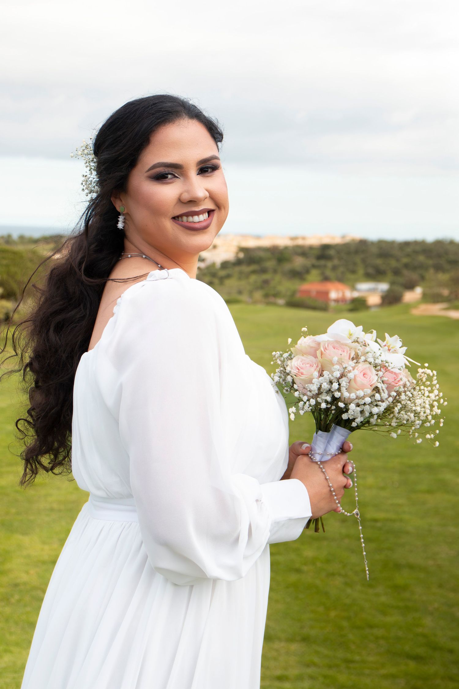 A bride in a flowy white dress poses with a delicate bouquet of pink and white flowers on a green golf course.