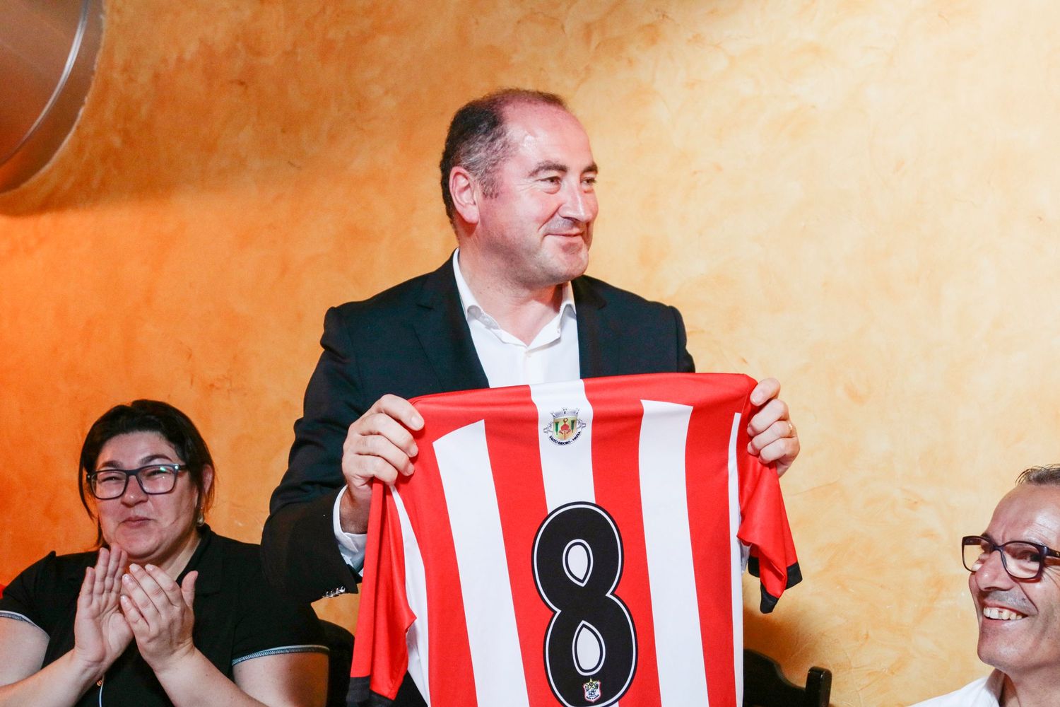 Someone holds up a red and white striped soccer jersey with number 8 against an orange wall as others look on.