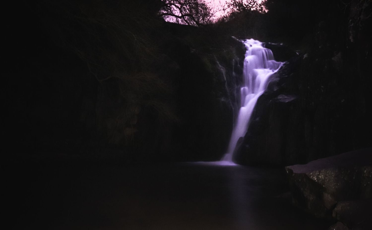 A majestic waterfall cascades down dark rocks at twilight creating a mystical purple and white scene in nature.