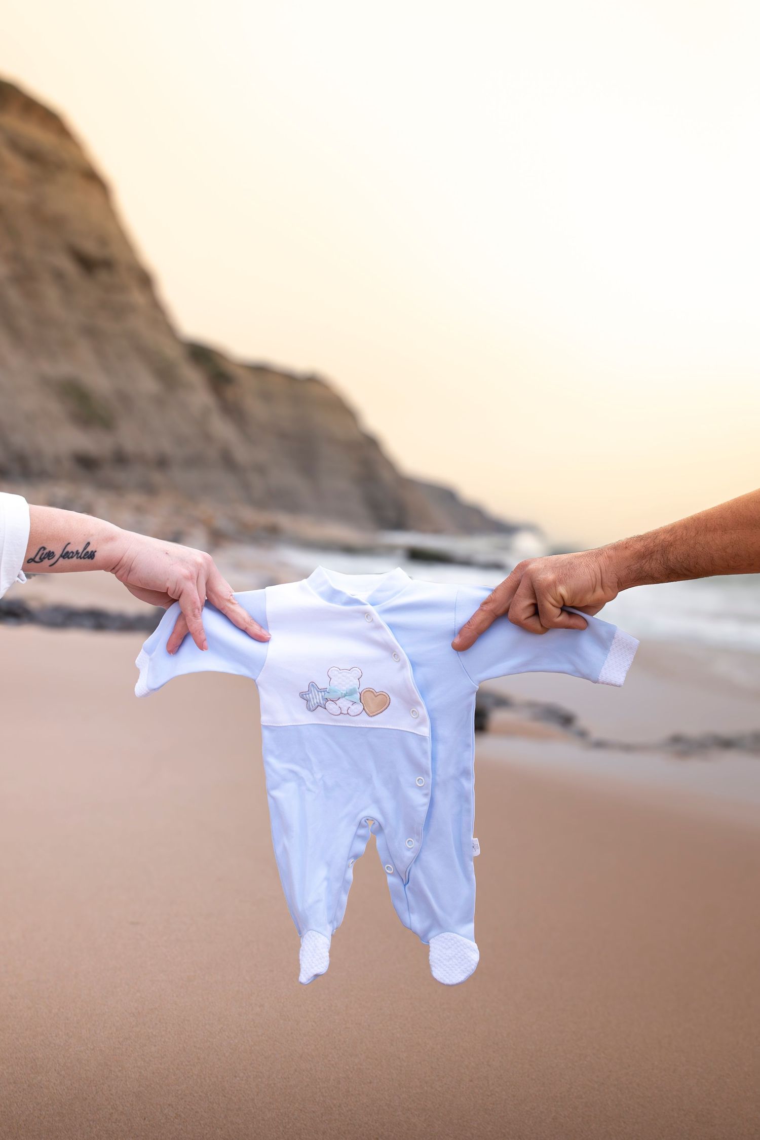 Two hands holding a white baby onesie on a sandy beach at sunset with rocky cliffs in the background.