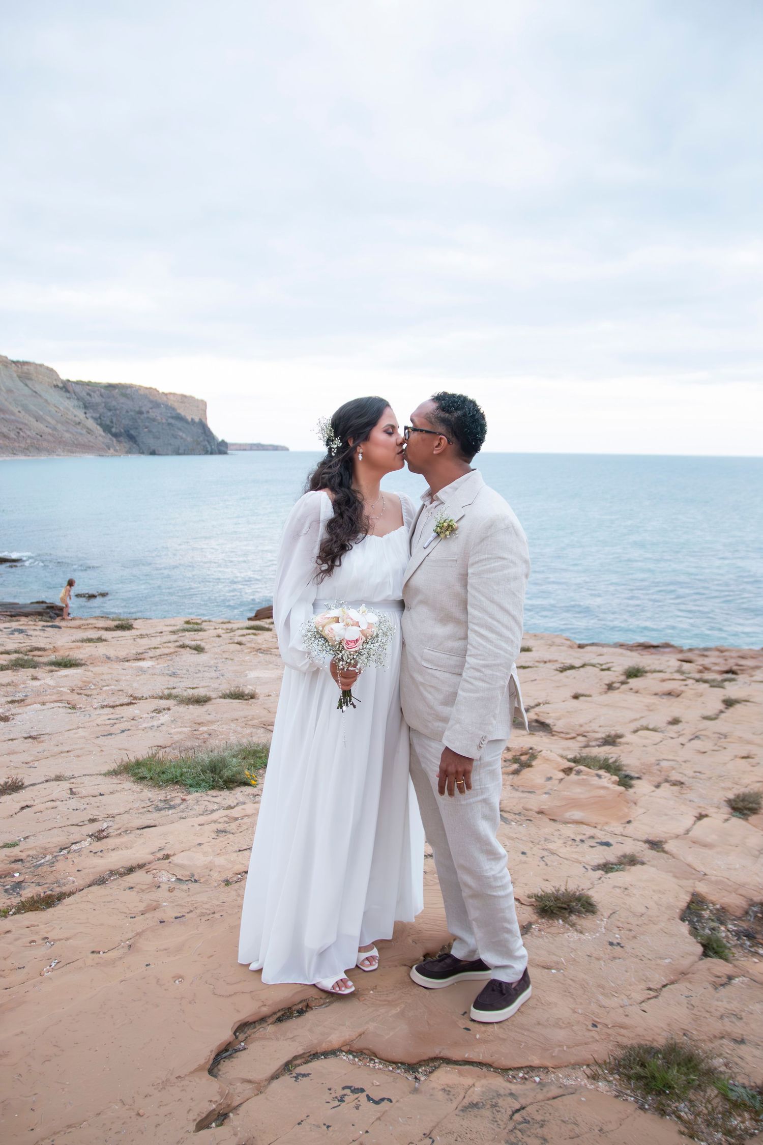 A newlywed couple embraces on a scenic rocky shoreline with ocean waves and cliffs in the background.