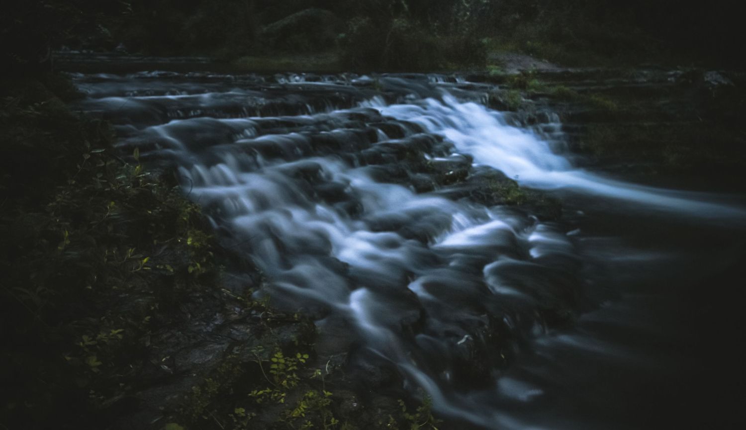 Long exposure photograph of cascading waterfall water flowing over dark rocks in low light conditions.