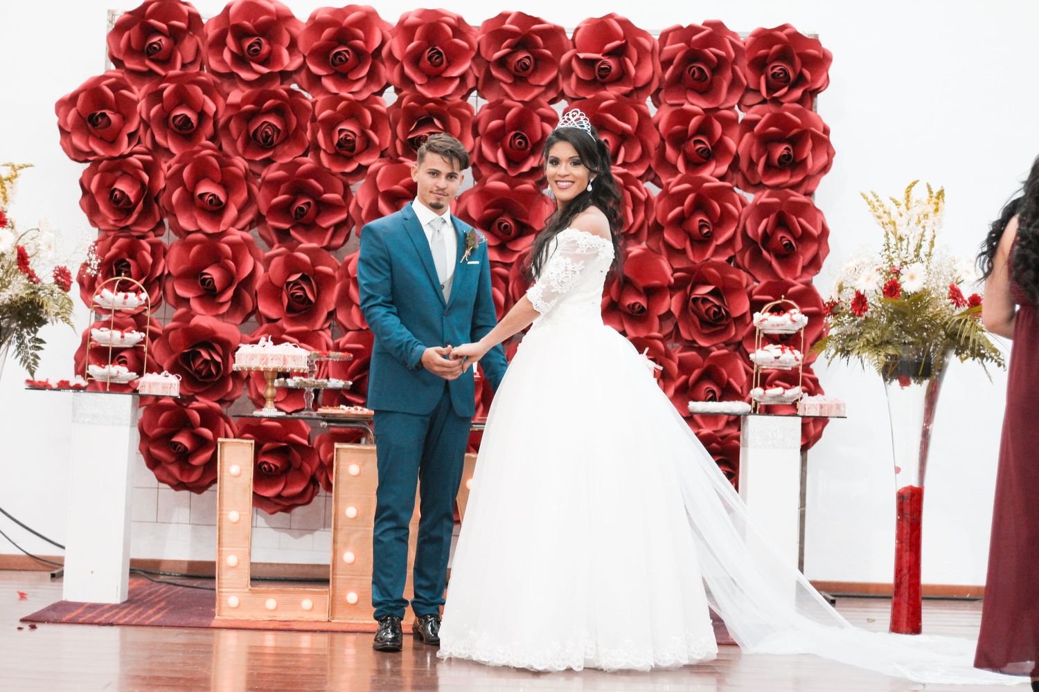A wedding couple holds hands in front of a dramatic wall of red roses and elegant white decor.