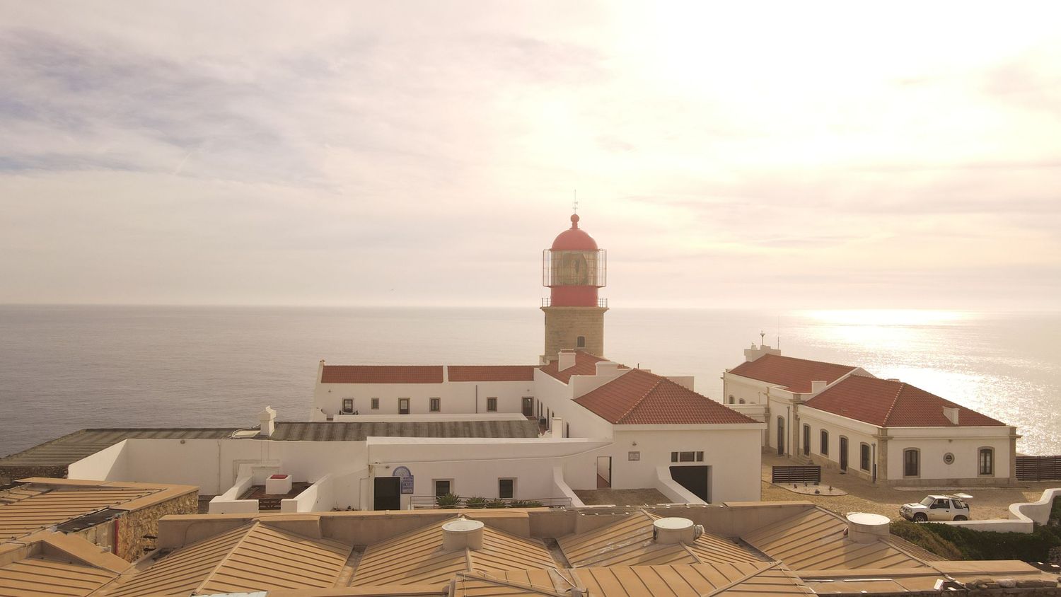Aerial view of historic lighthouse and white buildings along rugged coastline at sunset in Portugal.