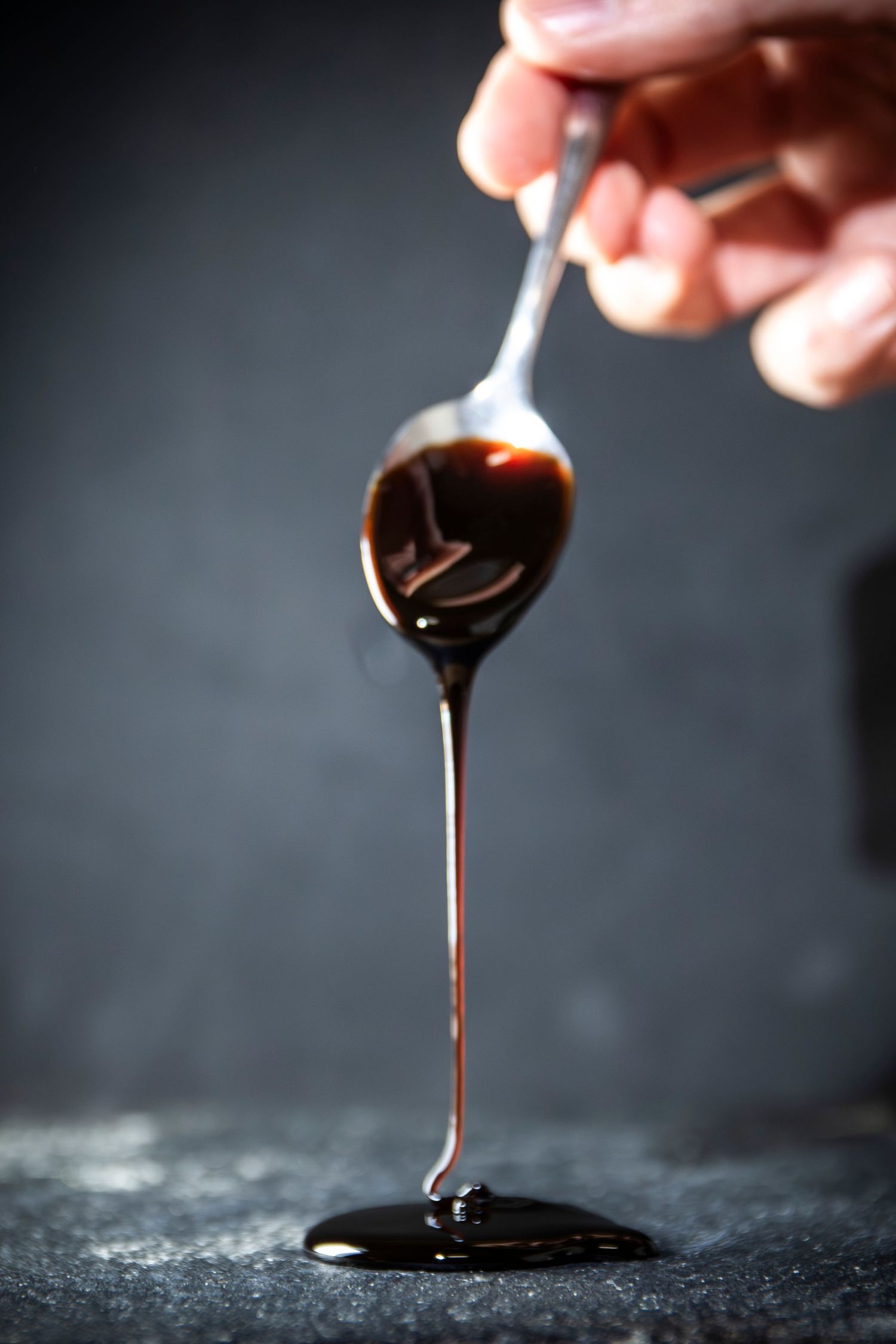 Dark syrupy liquid dripping from a spoon against a black background in dramatic lighting.