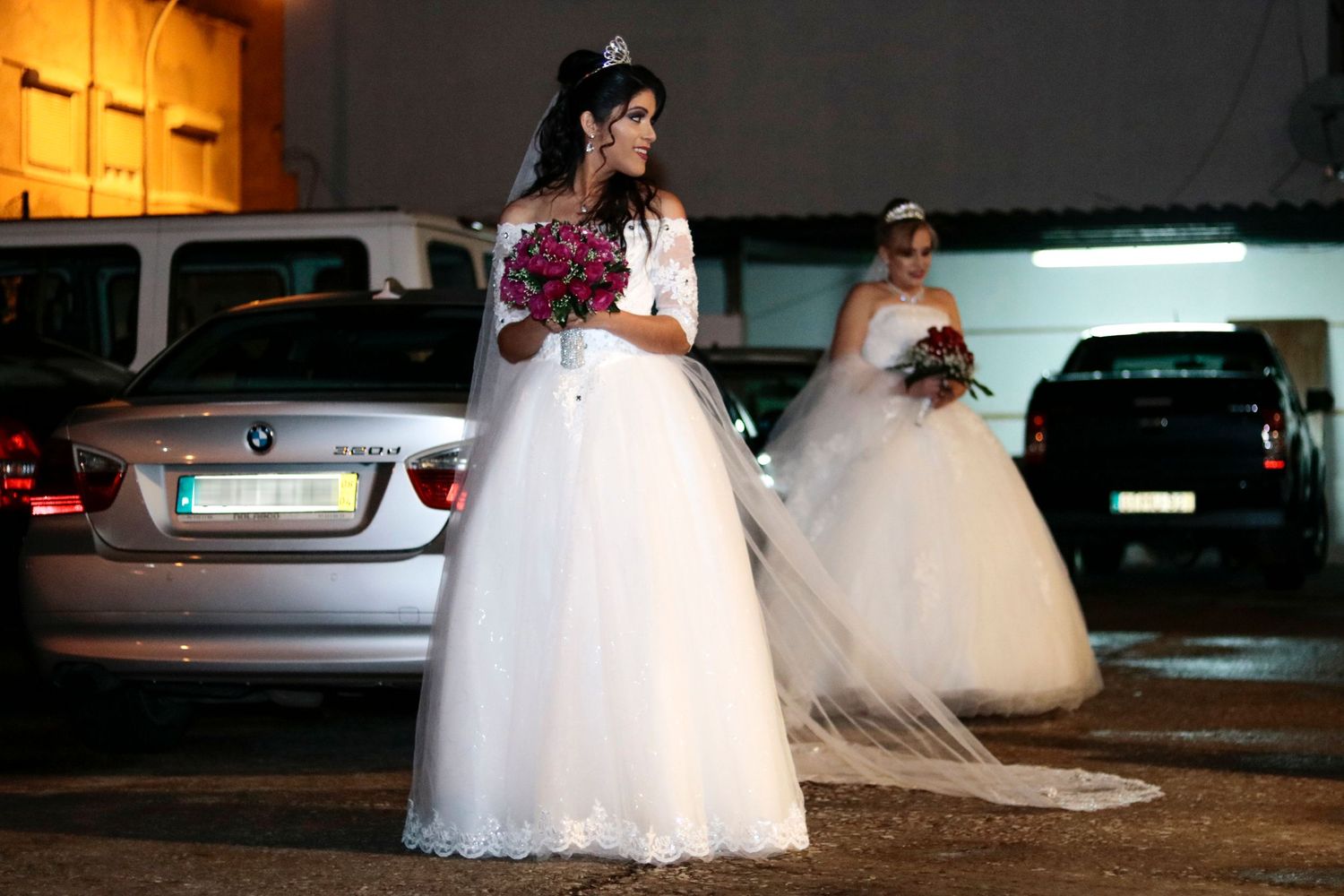 Two brides in elegant white wedding gowns stand in a dimly lit parking area at night.
