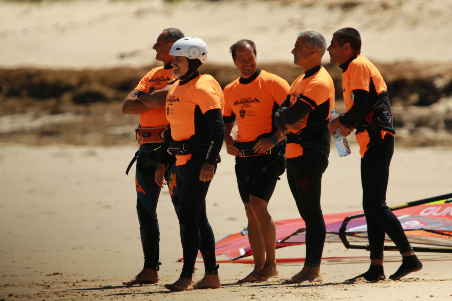 Group of windsurfers in orange uniforms gathering on the beach for instruction.