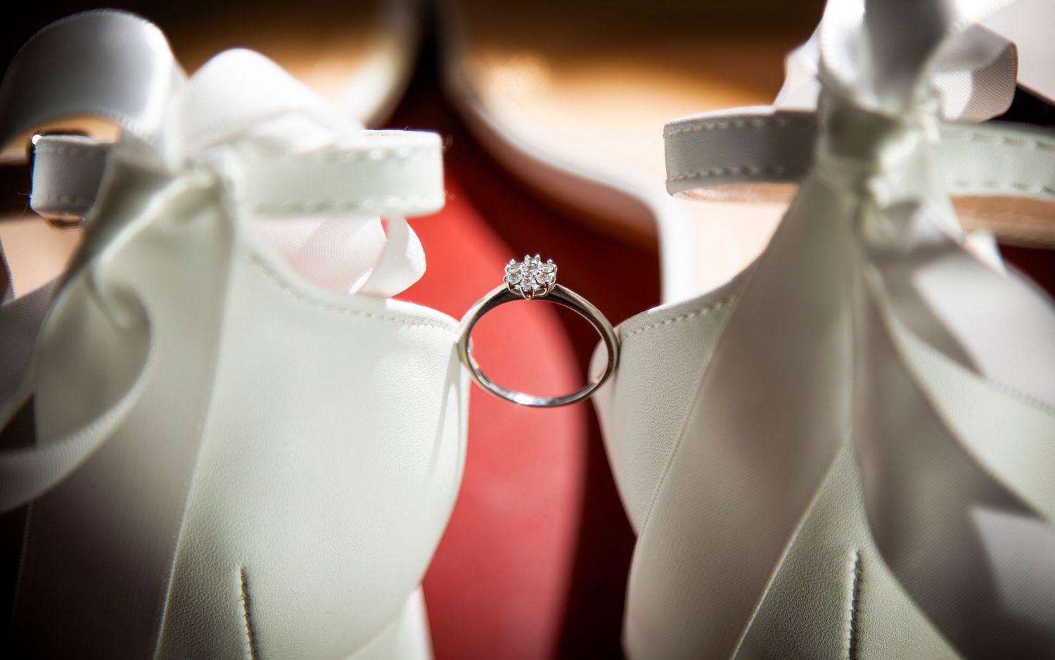A diamond ring artistically placed between two white bridal shoes.
