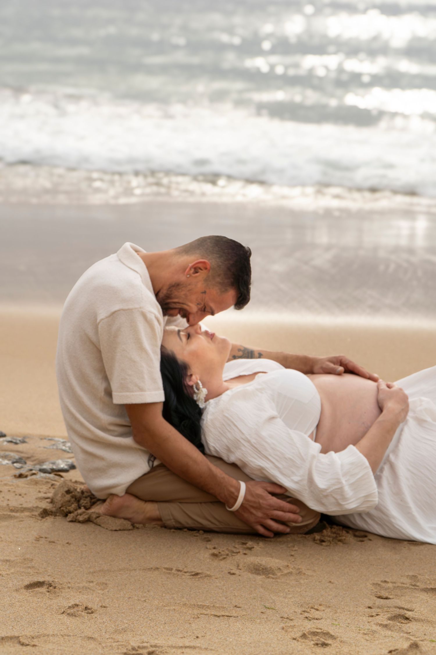 Pregnant couple sharing a tender moment on sandy beach, man leaning over woman lying down, ocean waves behind them.