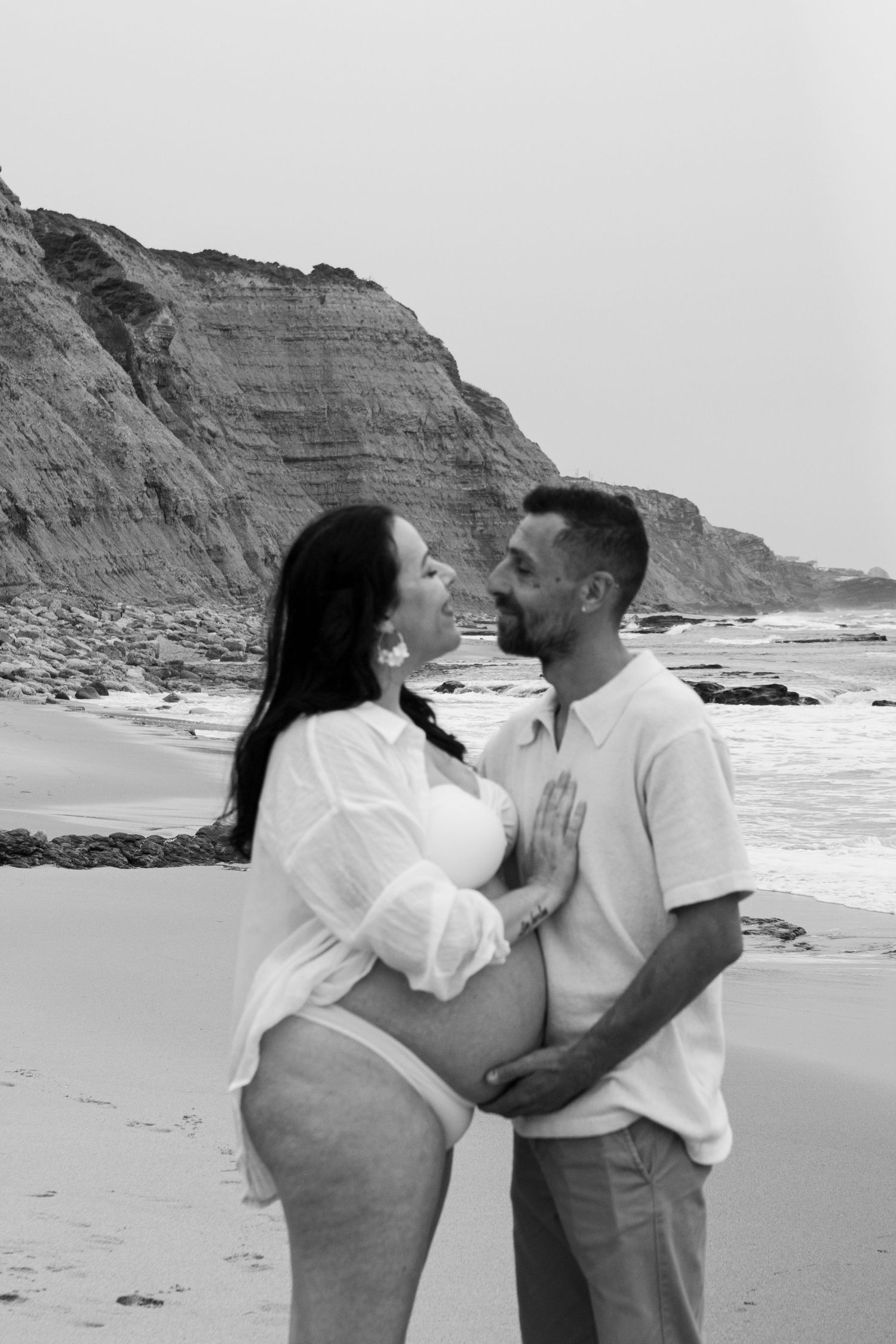 Black and white maternity photo of a couple embracing on a beach with dramatic cliffs in the background.