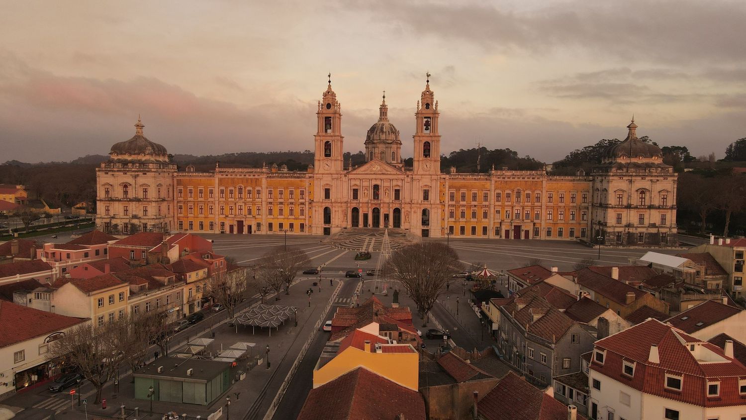 Aerial view of the ornate Mafra National Palace in Portugal illuminated during sunset with orange rooftops in foreground.
