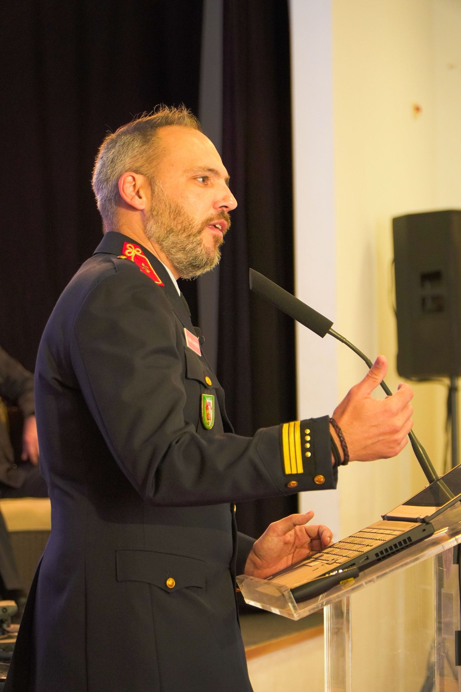 Speaker in military dress uniform speaks at podium during formal event with microphone in hand.